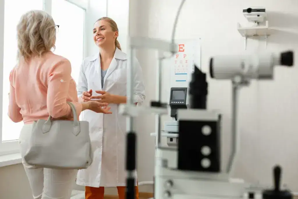 Optometrist in white coat smiling and consulting with a patient in a bright, welcoming eye care office, featuring eye examination equipment in the foreground.